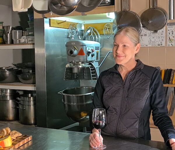 A woman stands behind a stainless steel bench in a commercial kitchen holding a glass of red wine and talking to another lady on the other side of the bench. Behind her someone films them on an phone.