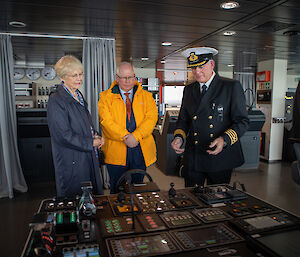 A man in a naval uniform explaining some instruments on a ship bridge to a man and a woman.