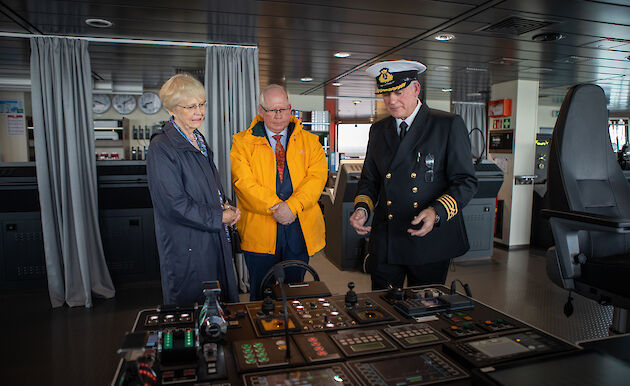 A man in a naval uniform explaining some instruments on a ship bridge to a man and a woman.