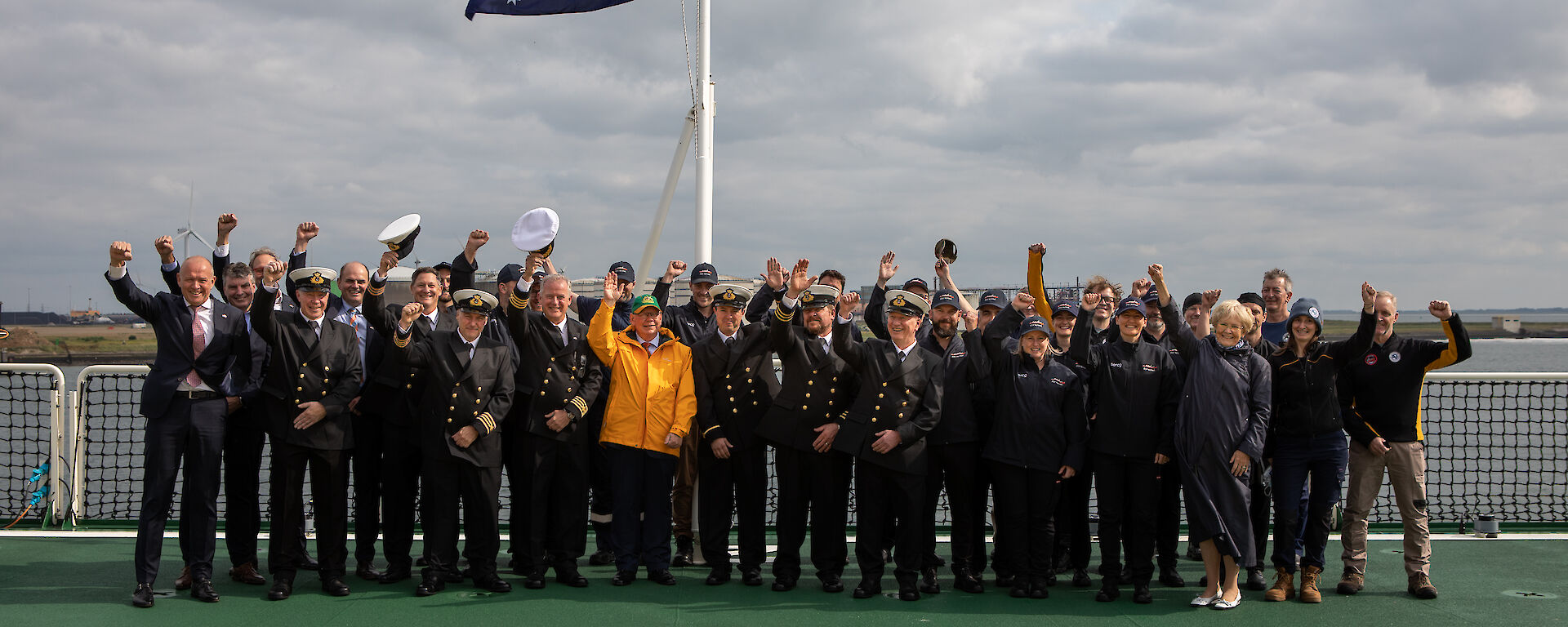 A large group of people standing on the deck of a ship underneath a flagpole with the Australian flag