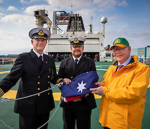 Three men on the deck of a ship centred round a folded up Australia flag smiling to camera.  Two men are in naval uniform.