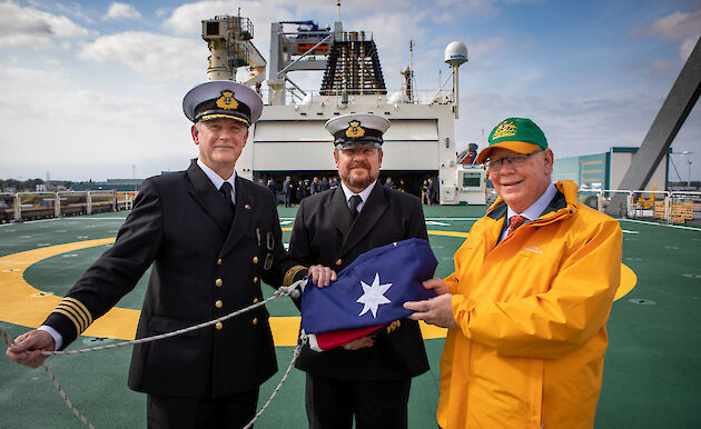 Three men on the deck of a ship centred round a folded up Australia flag smiling to camera.  Two men are in naval uniform.