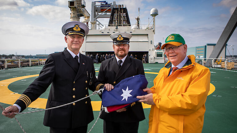 Three men on the deck of a ship centred round a folded up Australia flag smiling to camera. Two men are in naval uniform.