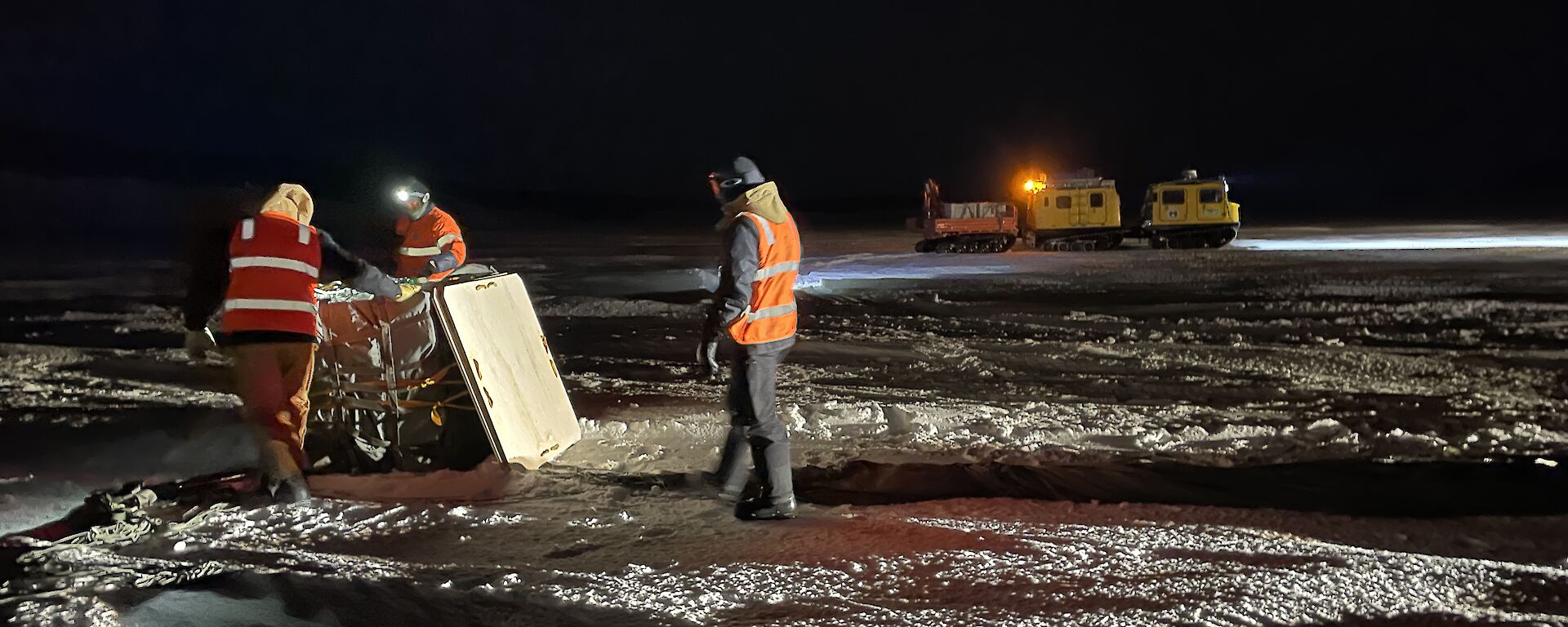 Three expeditioners in hi-visibility clothing standing around a cargo pallet with the yellow Hagglund in the background.