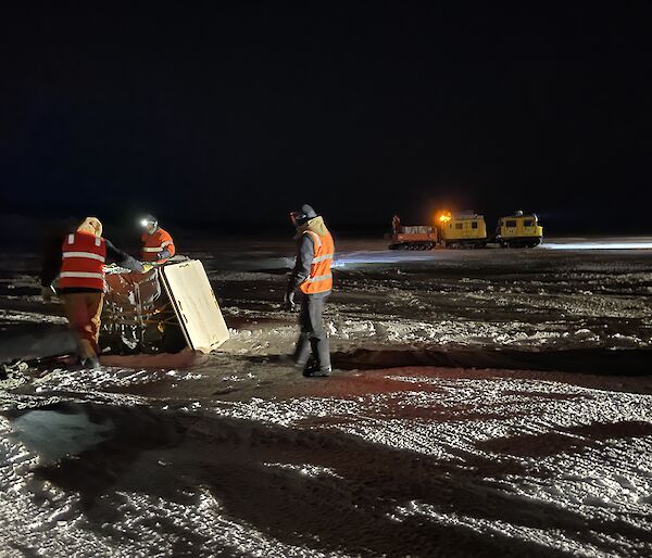 Three expeditioners in hi-visibility clothing standing around a cargo pallet with the yellow Hagglund in the background.