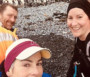 A selfie of three expeditioners on a pebble beach with king penguins in the background.