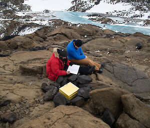 Two men sitting on some rocks looking at a log book, next to a cache box.