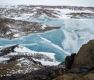 Looking down some rocky crags towards a frozen blue ice lake.