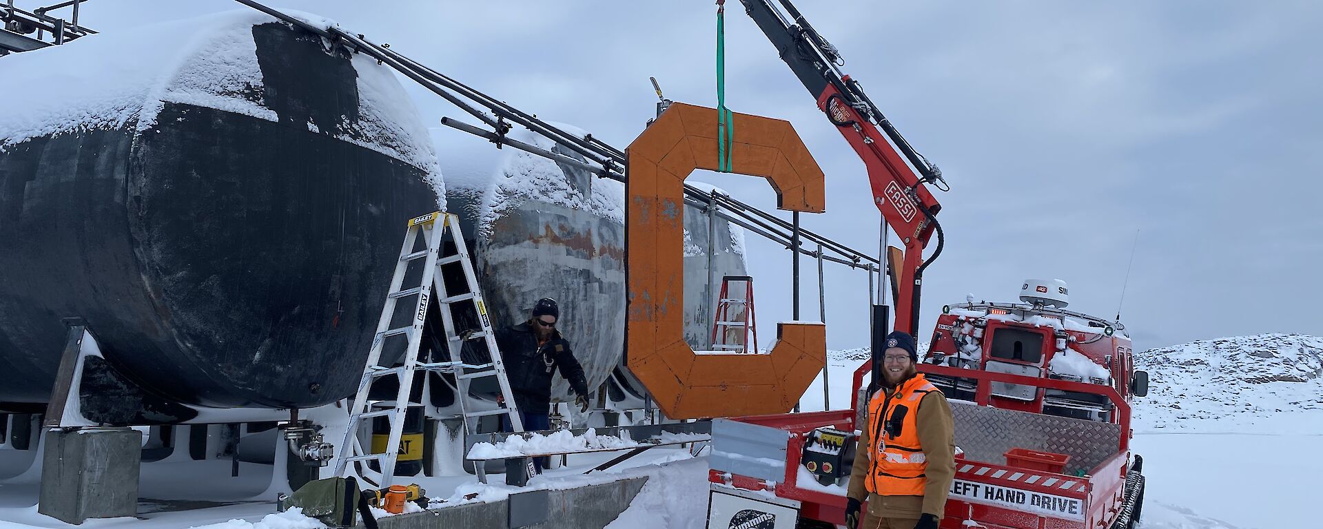 A man standing in the snow beside a truck with a small winch.  The winch is holding a large letter C .