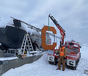 A man standing in the snow beside a truck with a small winch.  The winch is holding a large letter C .