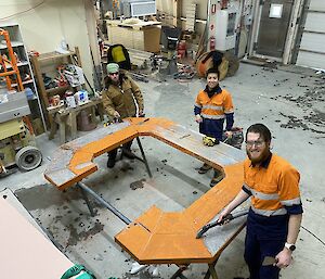 Three men in a workshop smiling to camera as they work on cleaning up a large metal letter C