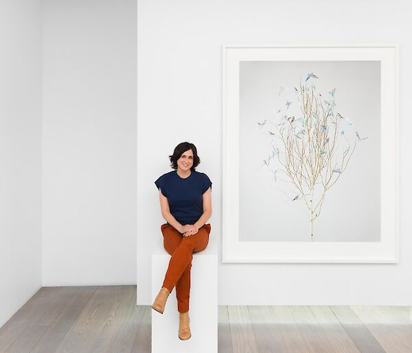 A lady sits on a white podium in an art gallery in front of a print of a tree filled with blue budgerigars