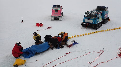Four scientists on the sea ice deploying instruments into a hole, with two Hägglunds vehicles behind.
