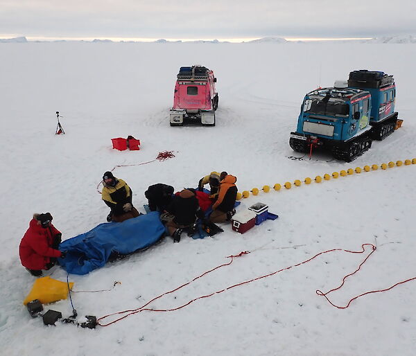 Four scientists on the sea ice deploying instruments into a hole, with two hagglund vehicles behind.