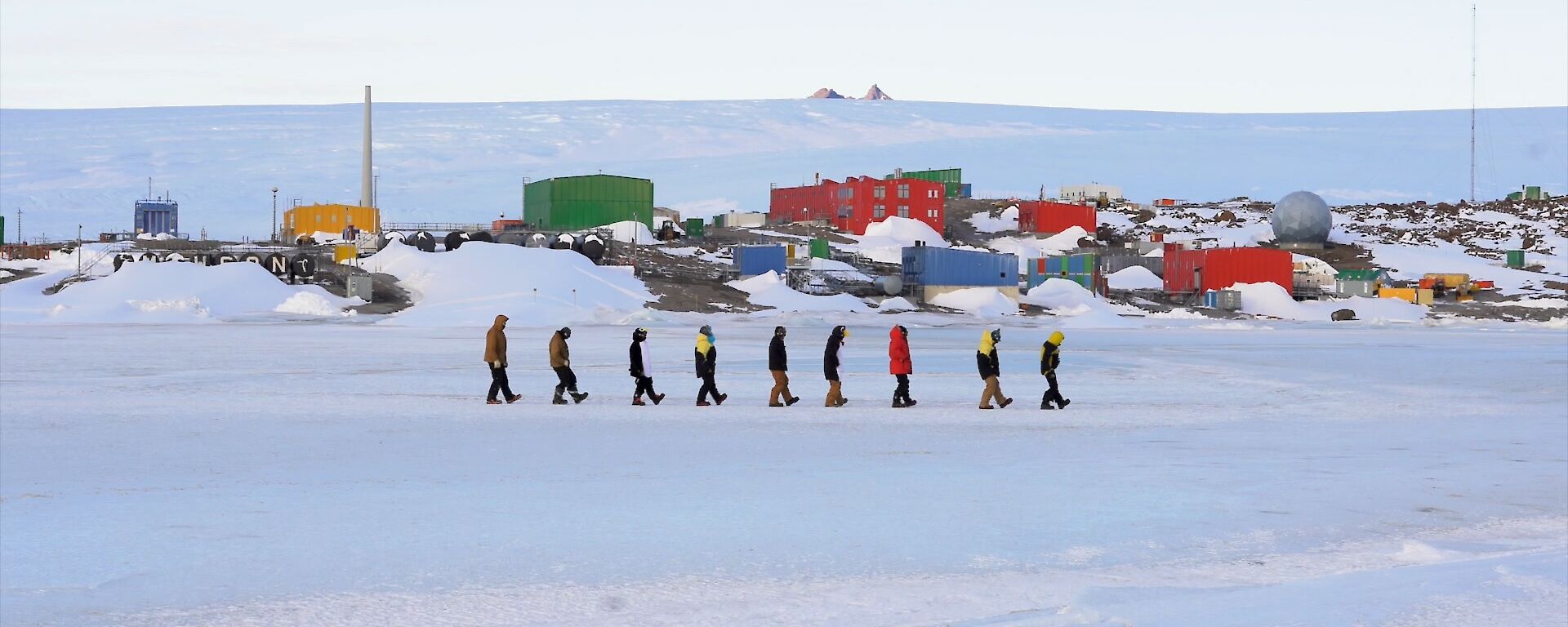 Nine expeditioners in a line boot scooting on the sea ice with Mawson station in the background.