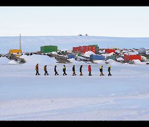 Nine expeditioners in a line boot scooting on the sea ice with Mawson station in the background.