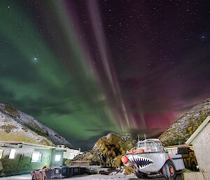 A couple of small sheds with a Larc (with painted teeth on it) parked outside in front of some hills.  A pink and green aurora is visible in the sky above.