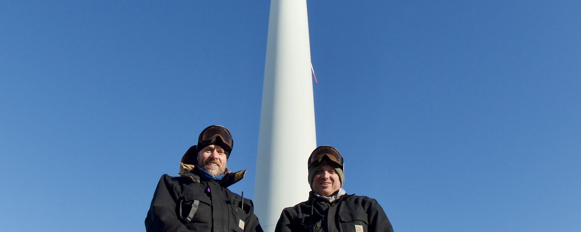 Two expeditioners standing for a photo with the wind turbine in the background.