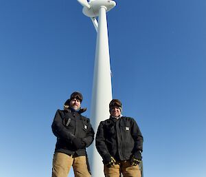 Two expeditioners standing for a photo with the wind turbine in the background.