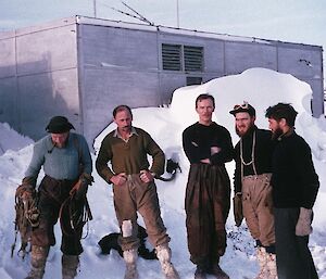 5 men stand in the snow in front of an original building - the first Davis station wintering party in 1957