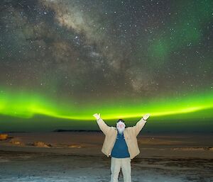 A man standing with his arms raised under a bright green winter aurora