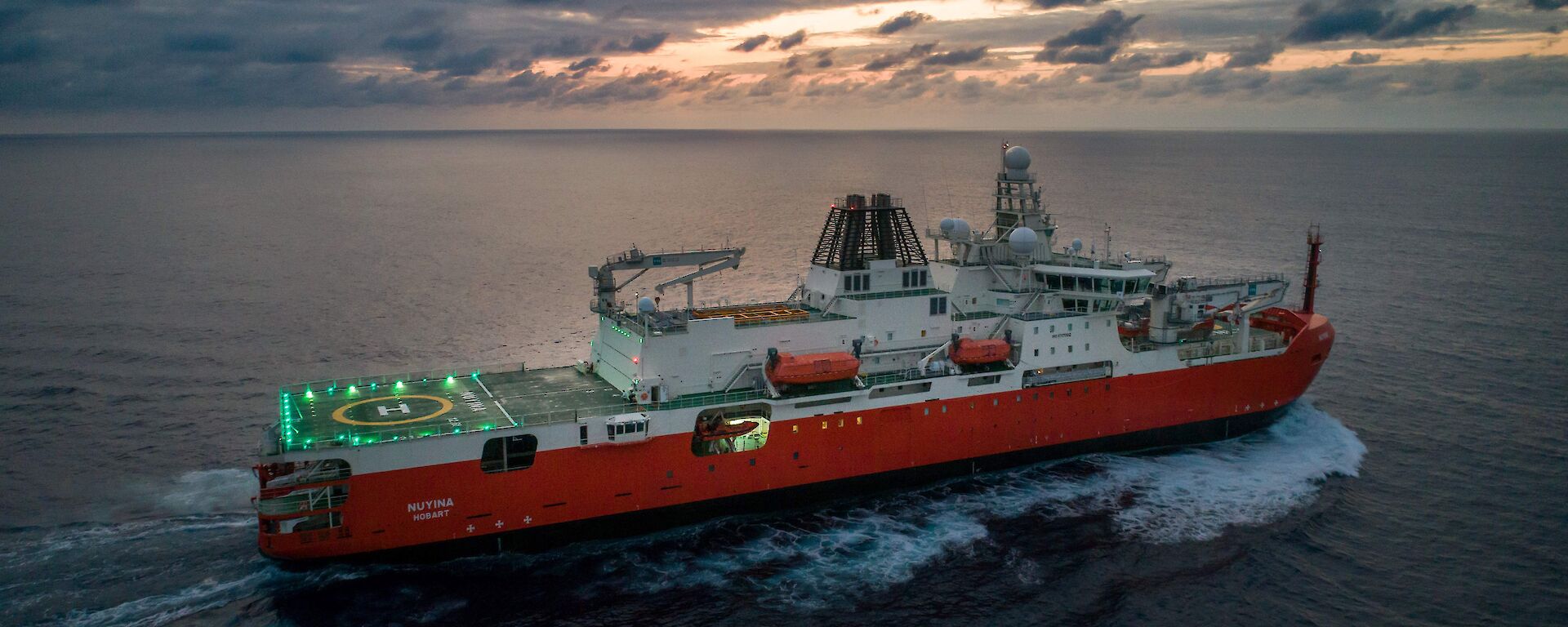 RSV Nuyina in the Atlantic Ocean at sunrise with lights illuminating the helideck.