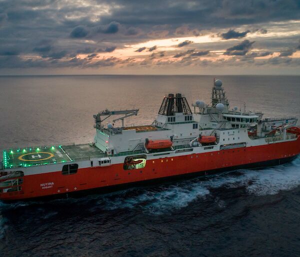 RSV Nuyina in the Atlantic Ocean at sunrise with lights illuminating the helideck.