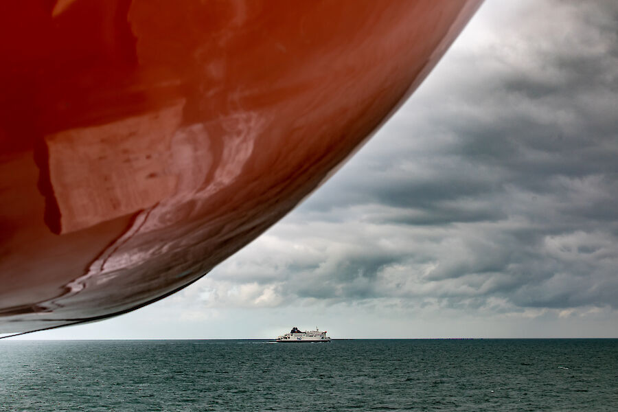 A large red ship bow covers the left of image and a small ferry on the horizon