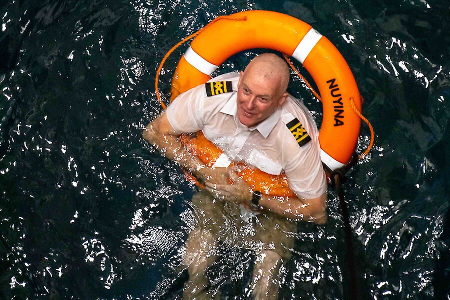 A man in a captains uniform and a life buoy over his head floating in some water