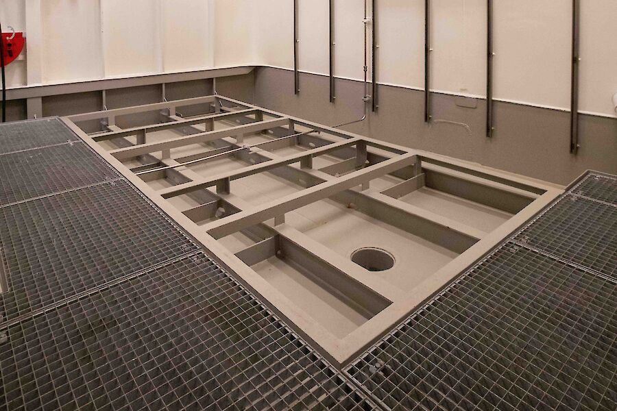 A room inside a ship with metal grids on the floor and a sunken area with a hole in it