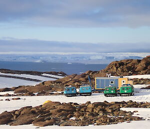 Two Hagglunds parked outside a field hut amongst a rocky and snow covered outcrop.