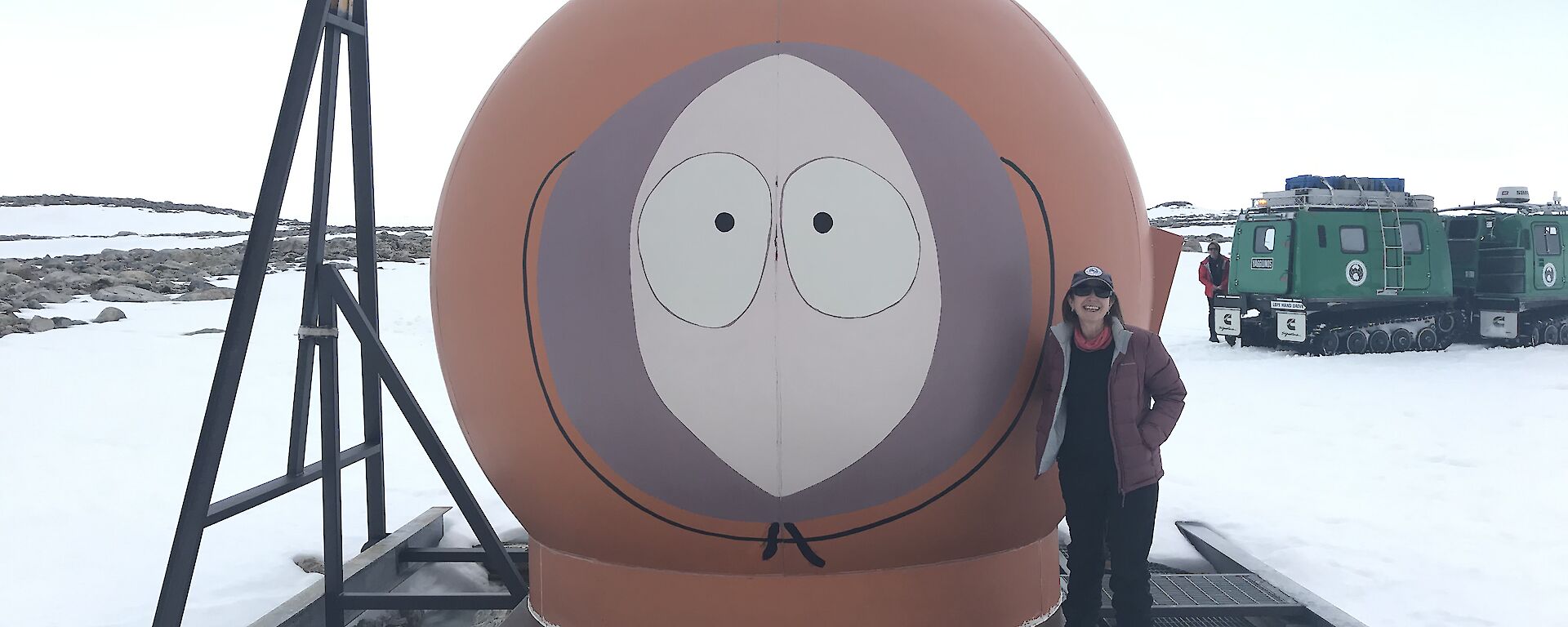A warmly clad person standing next to a round red hut that has a face on it (Kenny from Southpark).