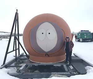 A warmly clad person standing next to a round red hut that has a face on it (Kenny from Southpark).