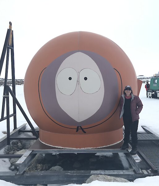 A warmly clad person standing next to a round red hut that has a face on it (Kenny from Southpark).