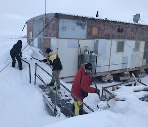 Three people with shovels moving snow away from a field hut.