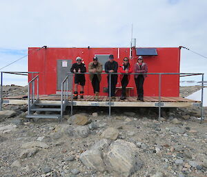 Five people stand in a row on the balcony of a red field hut on rocky ground.