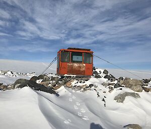 A small red toilet building tied down to a rocky outcrop with snow all around.