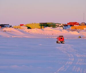 A Hagglund vehicle drives away from camera across the sea ice towards a cluster of colourful buildings in the snow