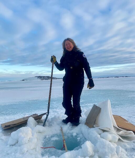 A woman stands next to a hole in the sea ice smiling to camera