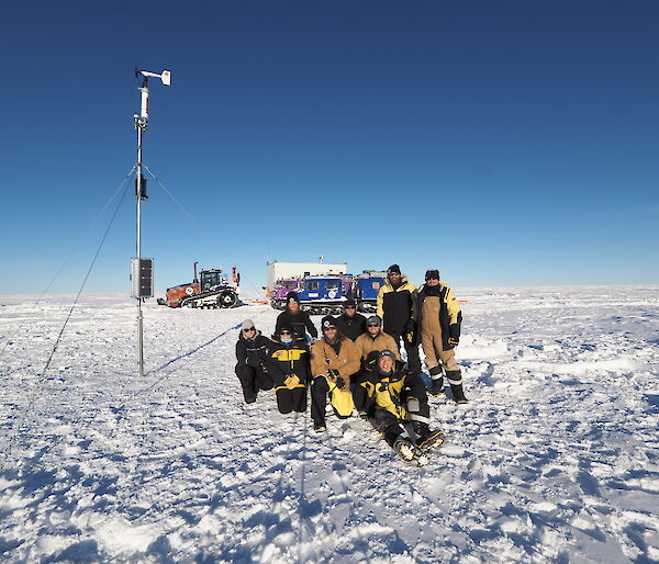 A group of expeditioners in yellow jackets sit in front of a tall Automatic Weather Station