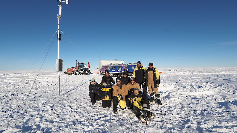 A group of expeditioners in yellow jackets sit in front of a tall Automatic Weather Station