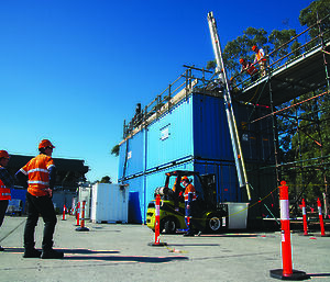 Four large blue containers stacked with scaffolding next to them.  Leaning on the containers is a long drill.  The work is being watched by several people in hi-vis