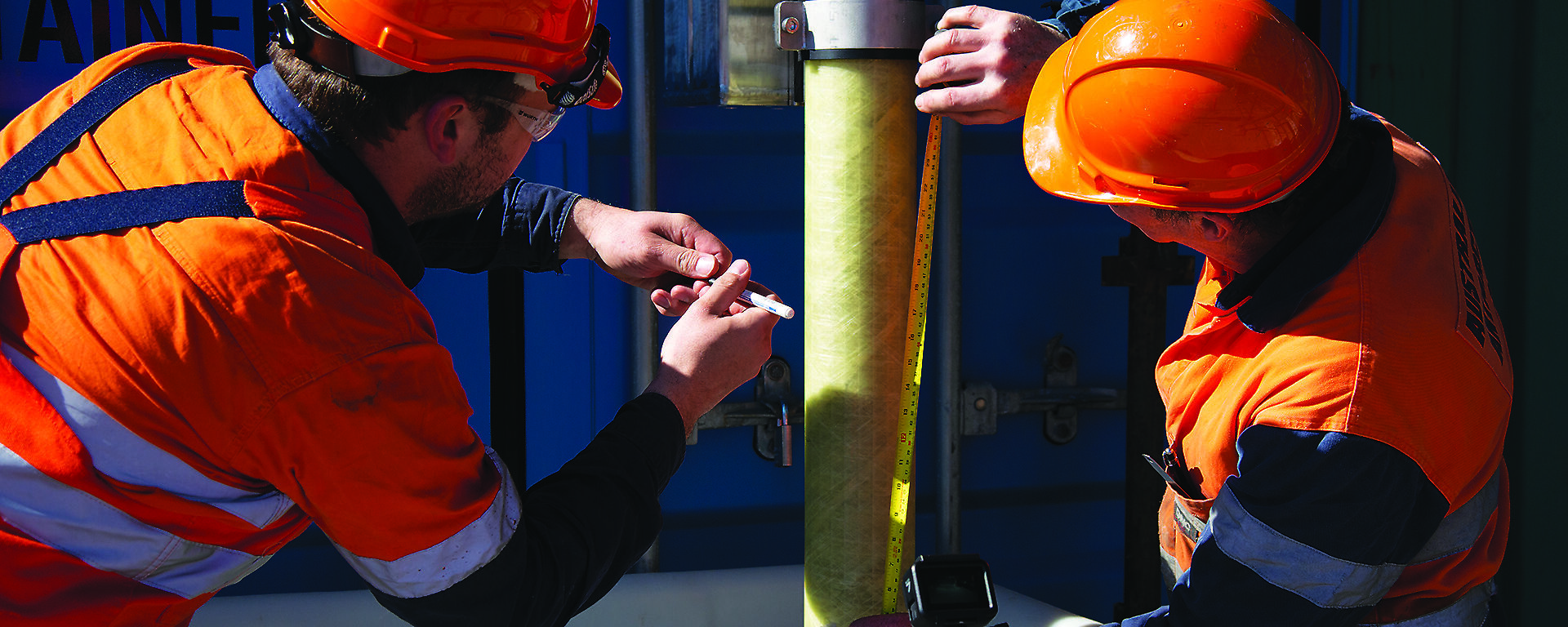 Two men in hi-vis and hard hats measuring the length of a drill pipe suspended over a block of ice