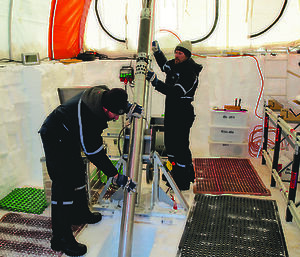Two men in a large tent built in to the ice,  manouvering a large drill in to a ice trench in the ground.