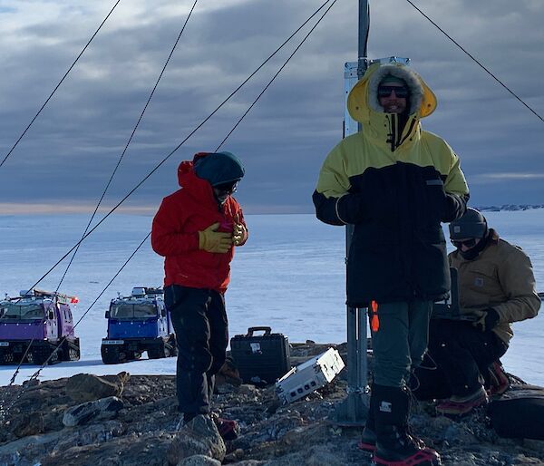 Three people stand next to an Automatic Weather Station with two large vehicles sitting on the snow behind them