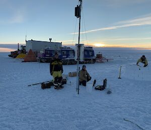 Two people working on a mast with vehicles and tents in the background