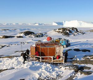 A field hut with expeditioners clearing snow off the walkway, the two Hagglund vehicles and an apple hut in the background with massive ice bergs trapped in the sea ice in the distance.