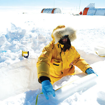 A scientist measuring an ice core with the Aurora Basin campsite in the background.