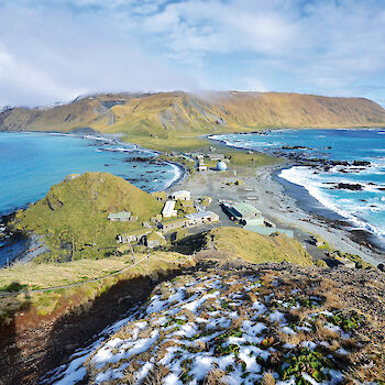 Elevated view on Macquarie Island looking south over narrow isthmus of land with station buildings to the island escarpment and plateau. Blue waters of bays on either side of isthmus.
