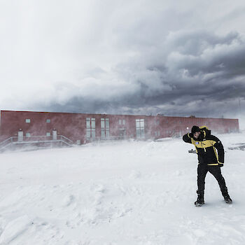 Expeditioner covering their face while bracing against strong wind and snow blow in front of station building.